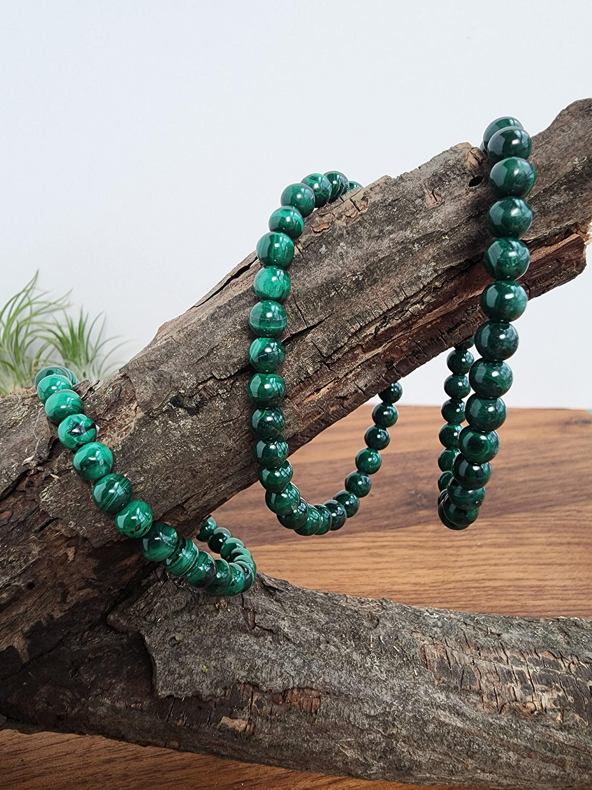 A necklace of green malachite beads is displayed on a wooden surface, with a plant visible in the background.
