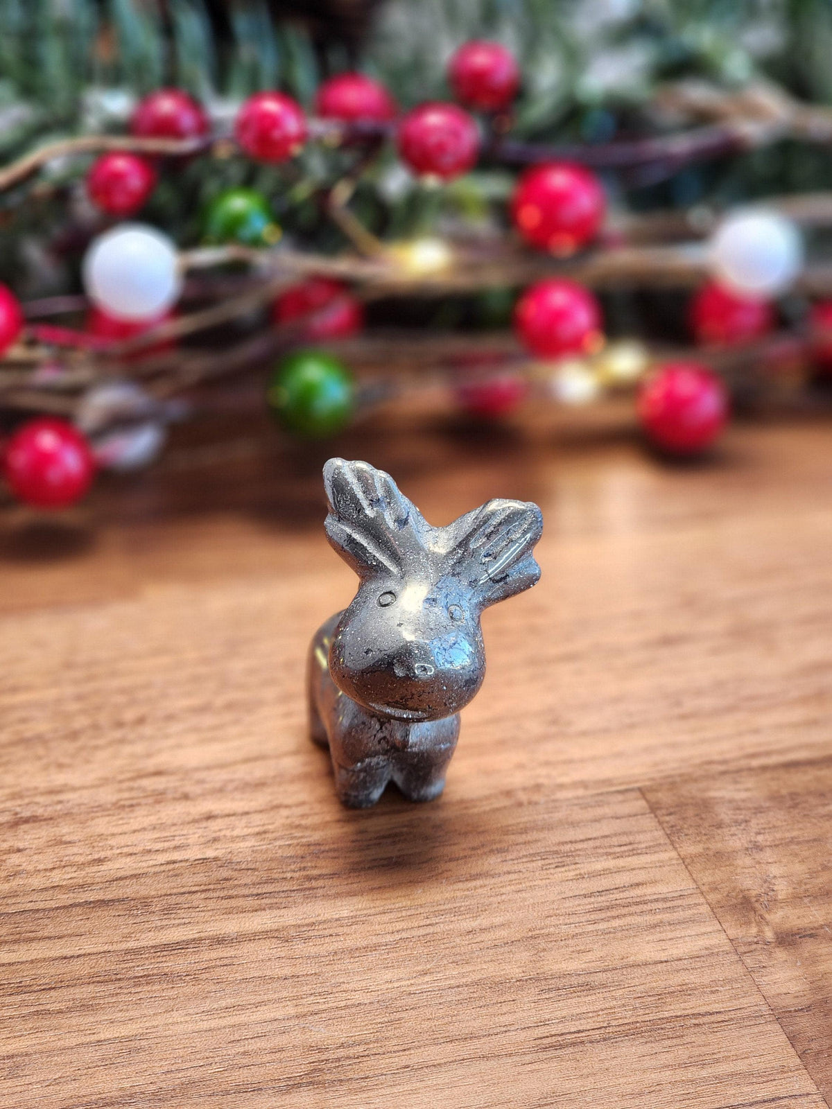 Mini Crystal Pyrite Reindeer with a tiny collar and adorable fluffy face carved into fools gold stands about 1.5" on a wooden tabletop near christmas lights, ferns and berries. Fully 3D hand carved golden moose.