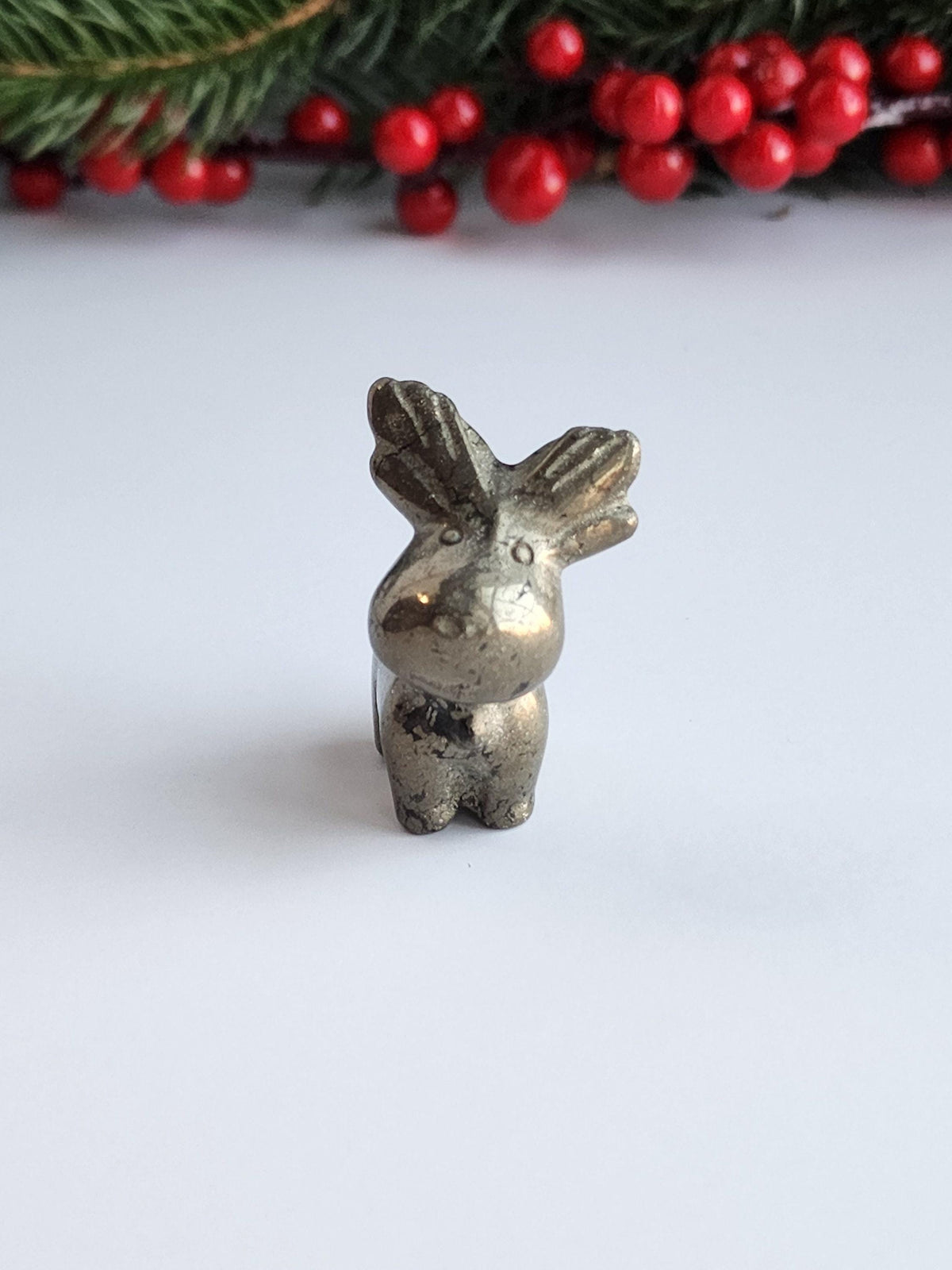 A small, shiny metal figurine of a reindeer is sitting on a white surface, with a sprig of greenery and red berries in the background.