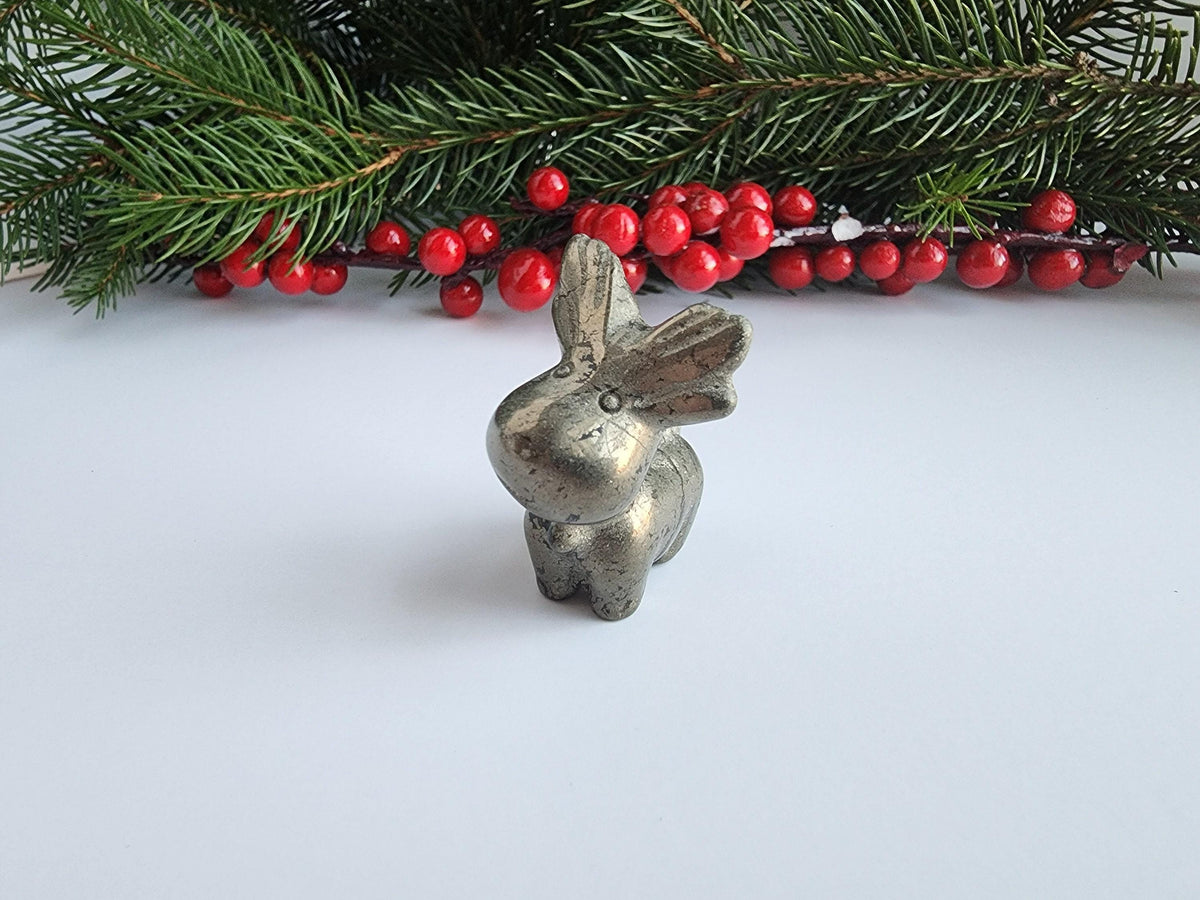 A small silver rabbit figurine is placed on a white surface, surrounded by red berries and pine branches.