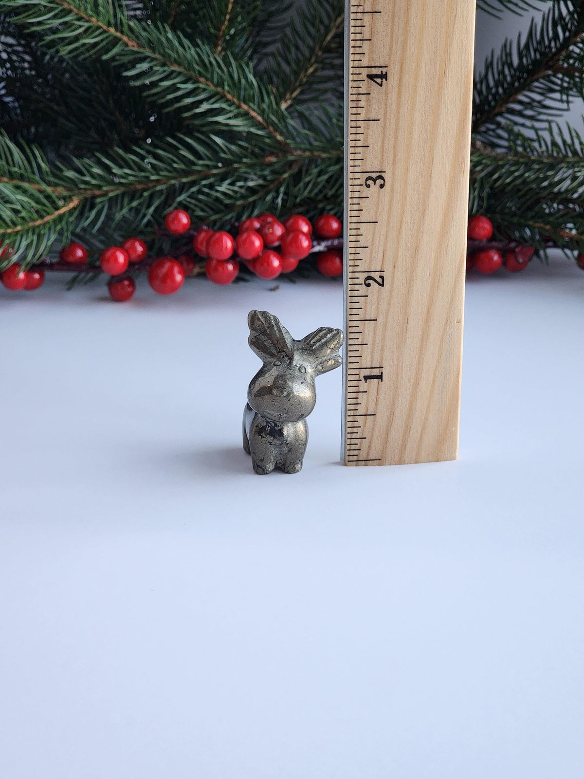 A small metal figurine of a reindeer is placed next to a ruler on a white surface, with a sprig of pine needles and red berries in the background.
