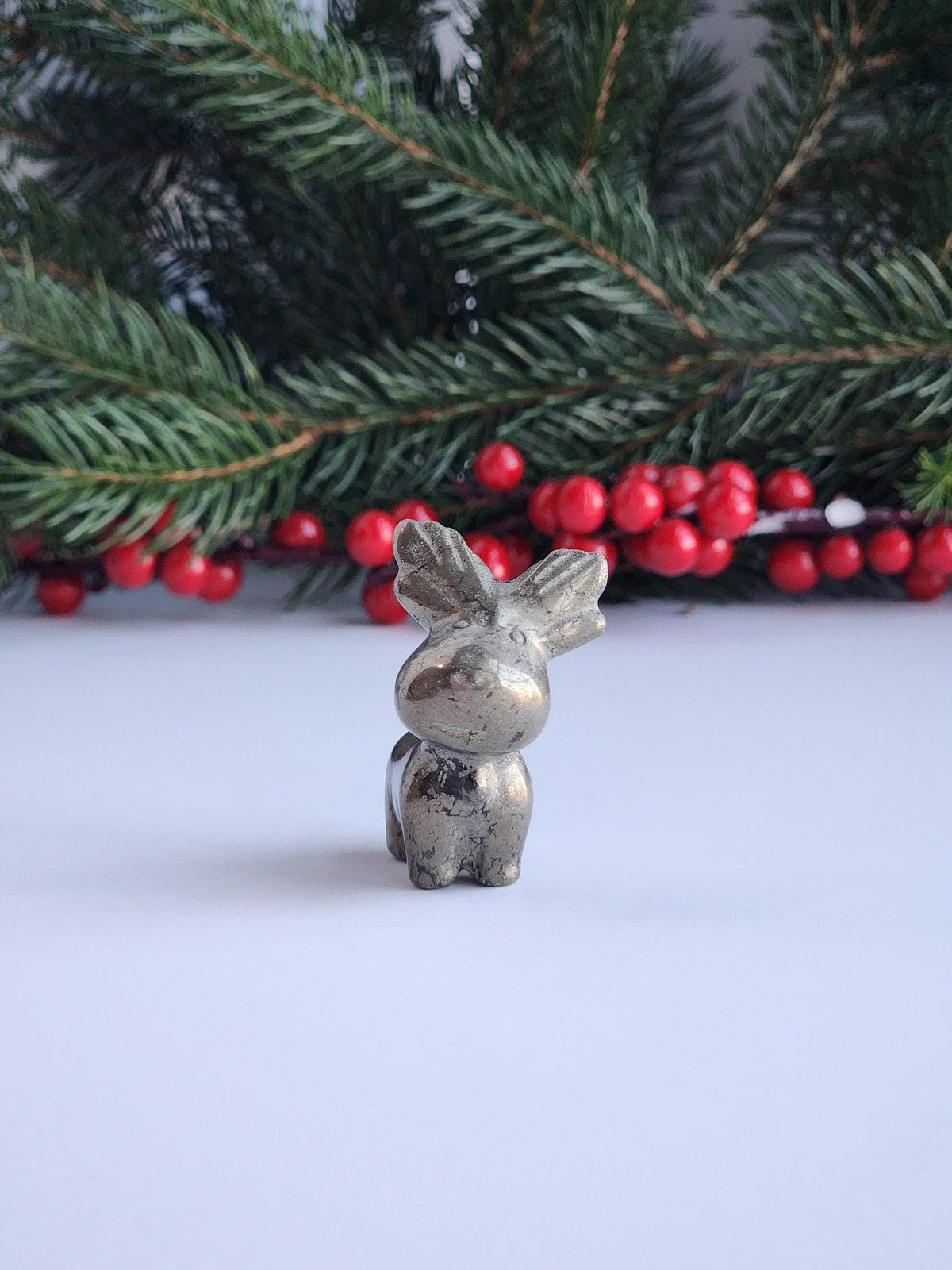 A small metal figurine of a reindeer is placed in front of a Christmas tree with red berries.
