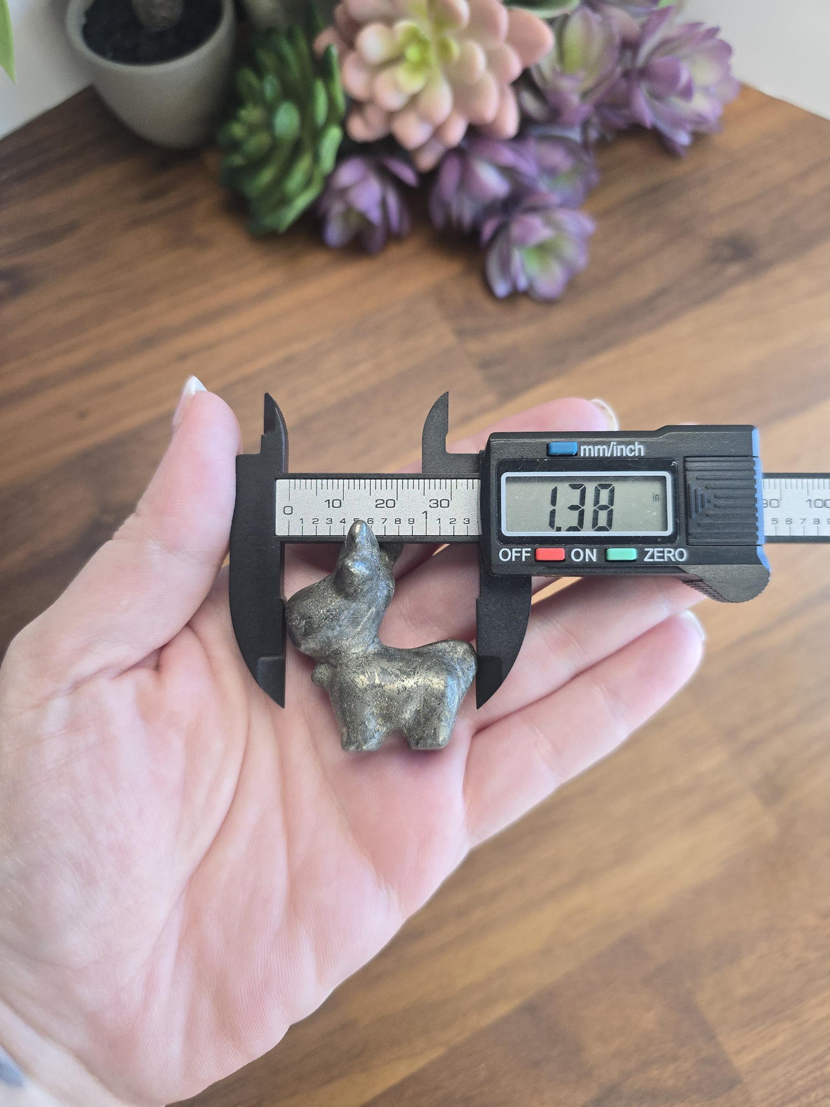 A person's hand holding a small metal object, possibly a toy or figurine, next to a digital caliper that measures the object's dimensions.