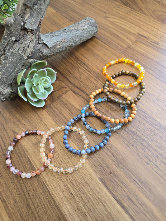 May include: A collection of eight stretch bracelets made from various round beads, including translucent orange, brown wood, light blue, dark grey, clear, and multi-colored stone beads, arranged in a cascading line on a wooden surface.