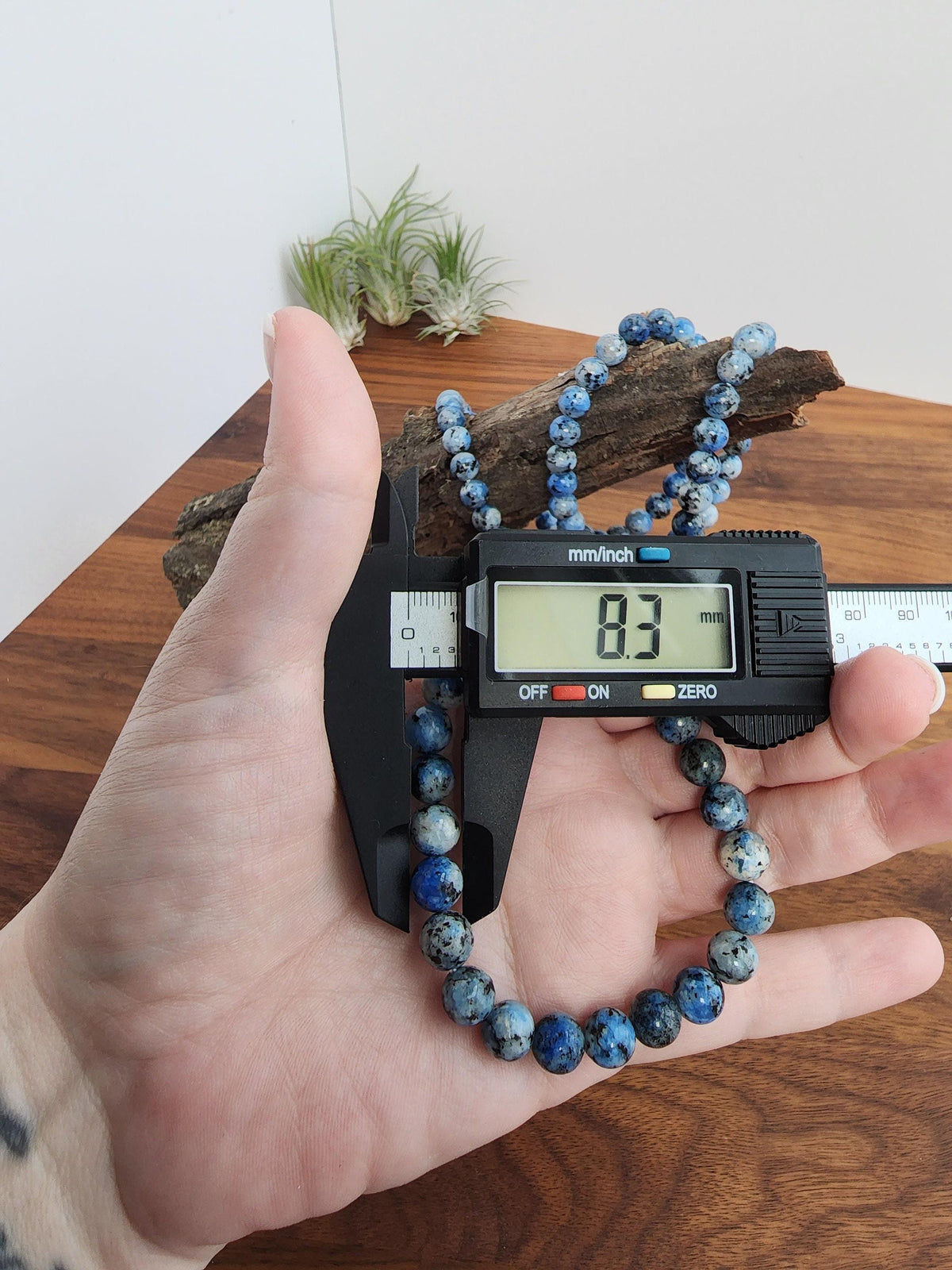 A person's hand holding a digital caliper to measure the size of a blue and white beaded necklace.
